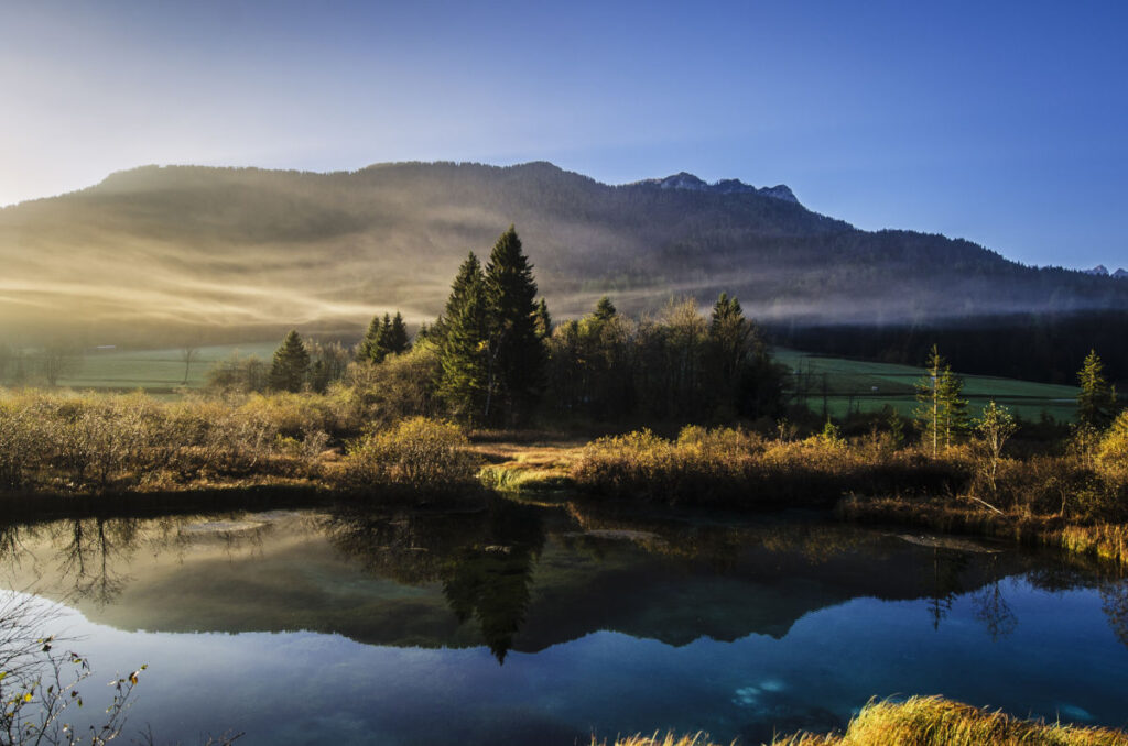 Image across a natural landsacpe with water in the foreground, then trees and low-hanging clouds, the hills and mountains in the back.